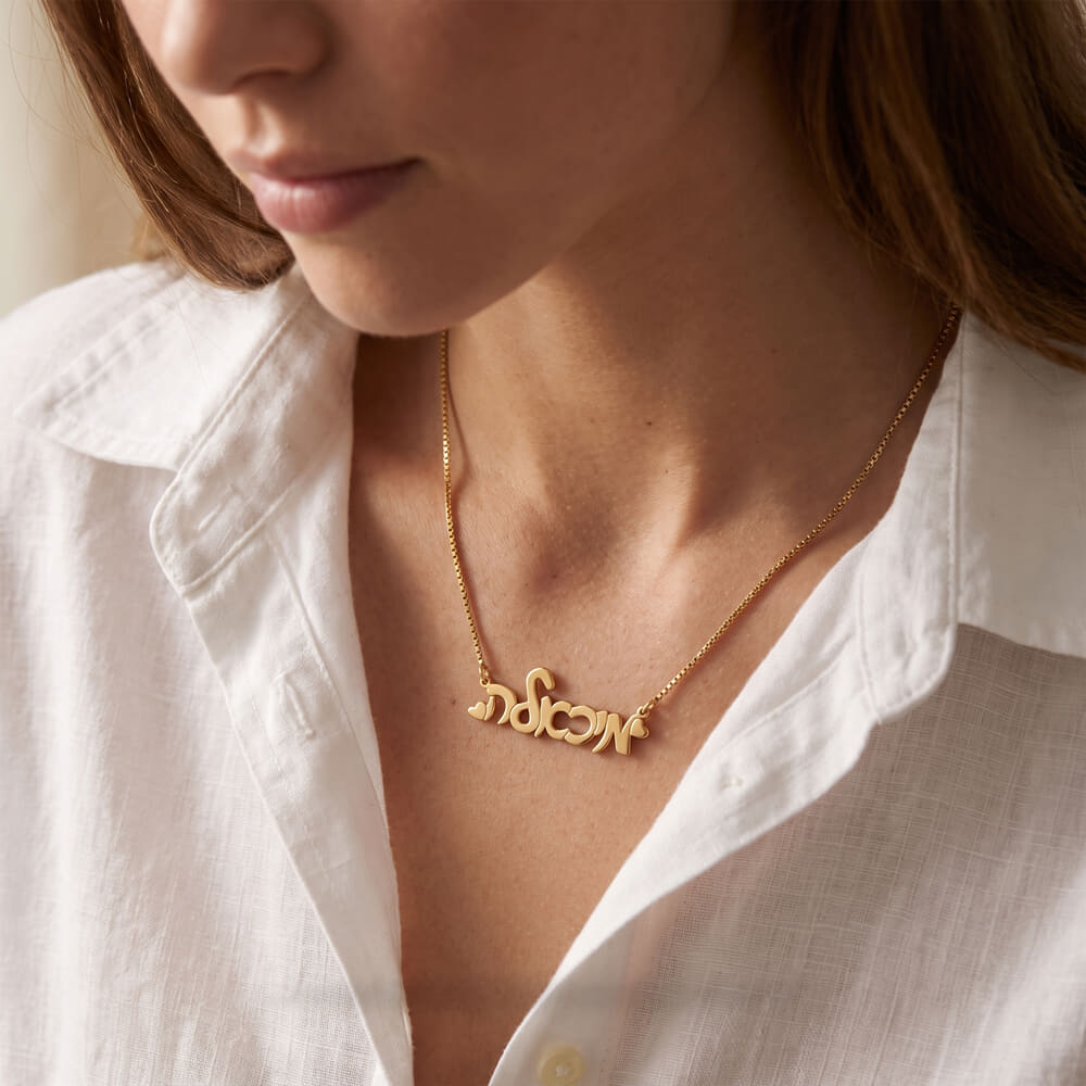 Woman elegantly wearing a cursive Hebrew name necklace with hearts, complementing her casual outfit